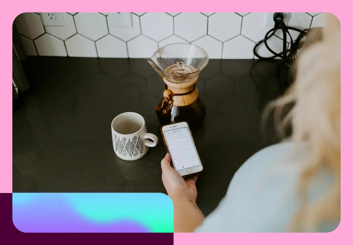 A woman holding her phone at a kitchen counter, next to a coffee mug and coffee maker