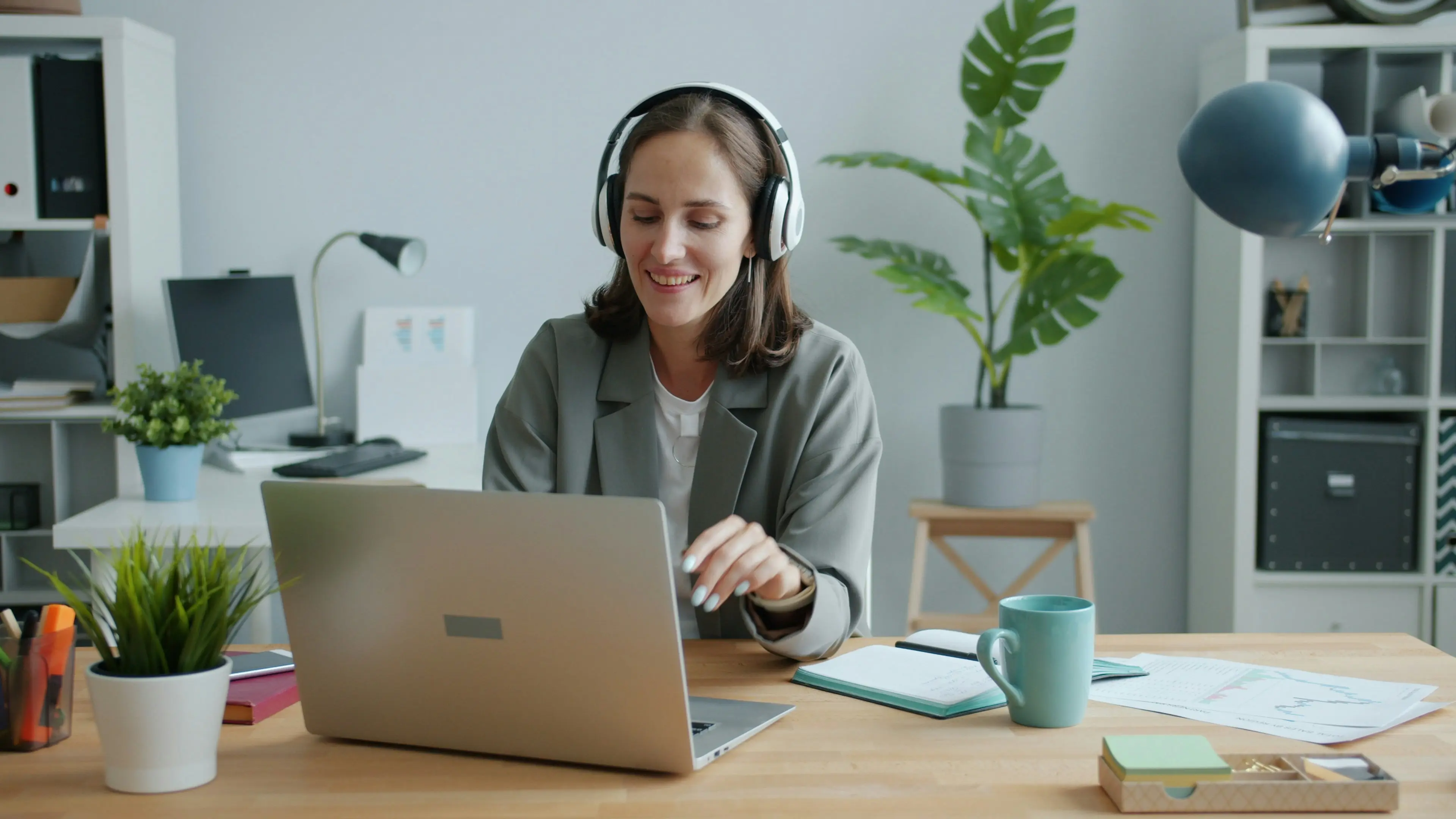 A woman wearing headphones smiles while typing on a laptop at her desk